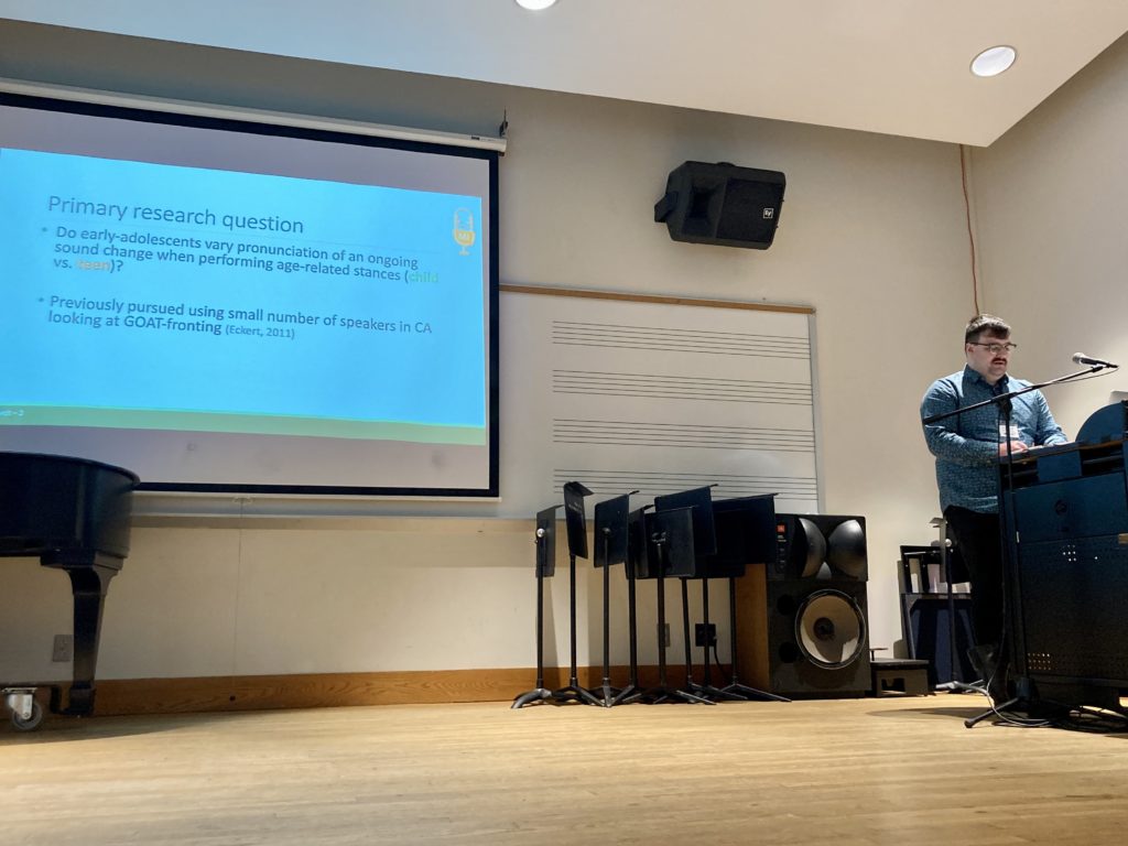 Adam Barnhardt stands behind a podium. A screen on the wall displays a slide titled "Primary research question." Below are two bullets. The first: "Do early adolescents vary pronunciation of an ongoing sound change when performing age-related stance (child vs teen)? Second: "Previously pursued using small number of speakers in CA looking at GOAT-fronting (Eckert 2011)"