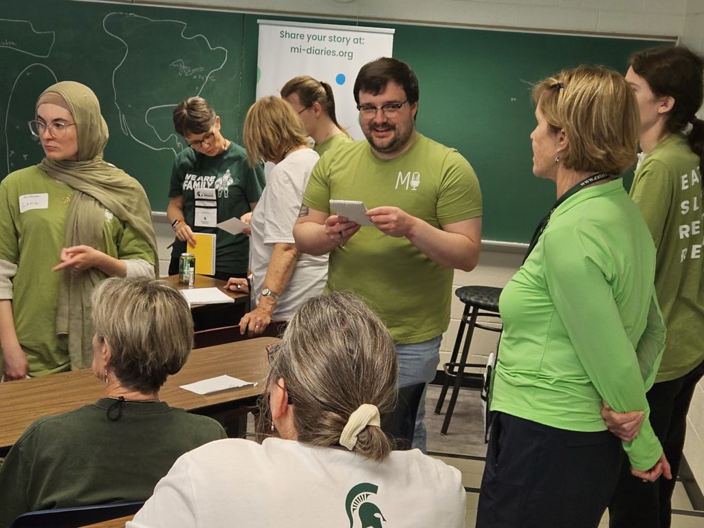 A photo taken in an MSU classroom. MI Diaries team members in green t-shirts are standing with and talking to grandparent participants around some tables.