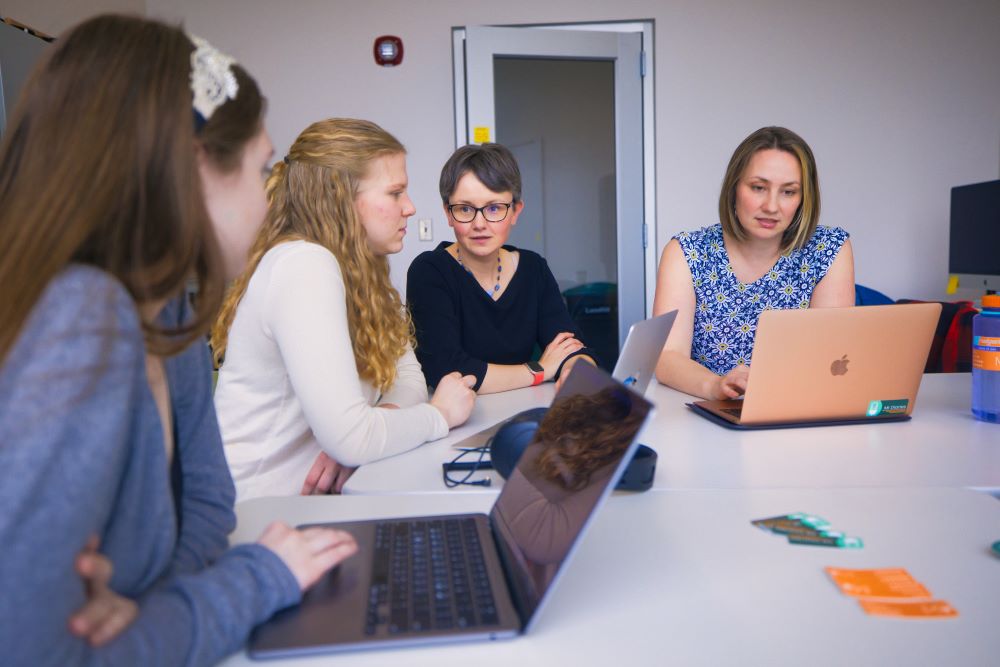 Photo of two students and two professors seated at a table. People are either talking to each other or looking at their open laptop.