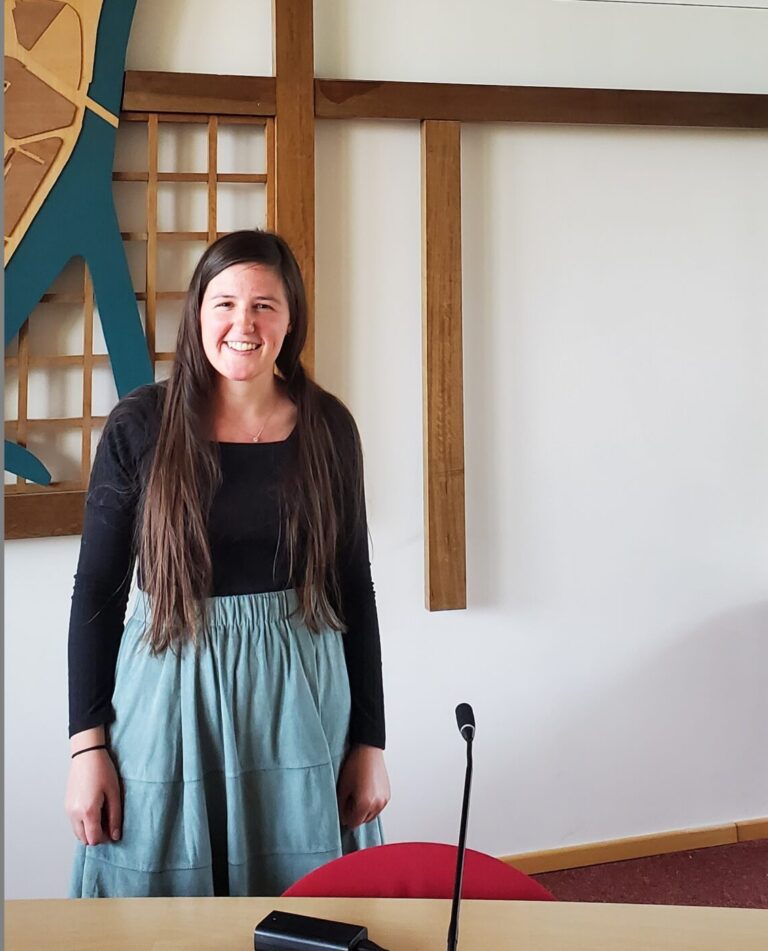 A photo of Rose Fisher smiling and standing in front of a table and a microphone.